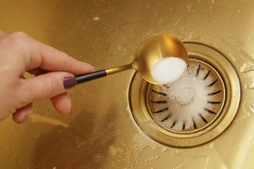 Hand holding a teaspoon of baking soda that's being poured into a sink drain.