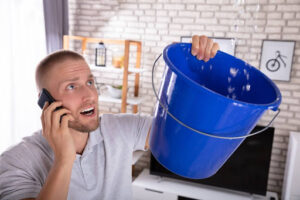 A man on the phone as he uses a bucket to catch water leaking from the ceiling in his home.